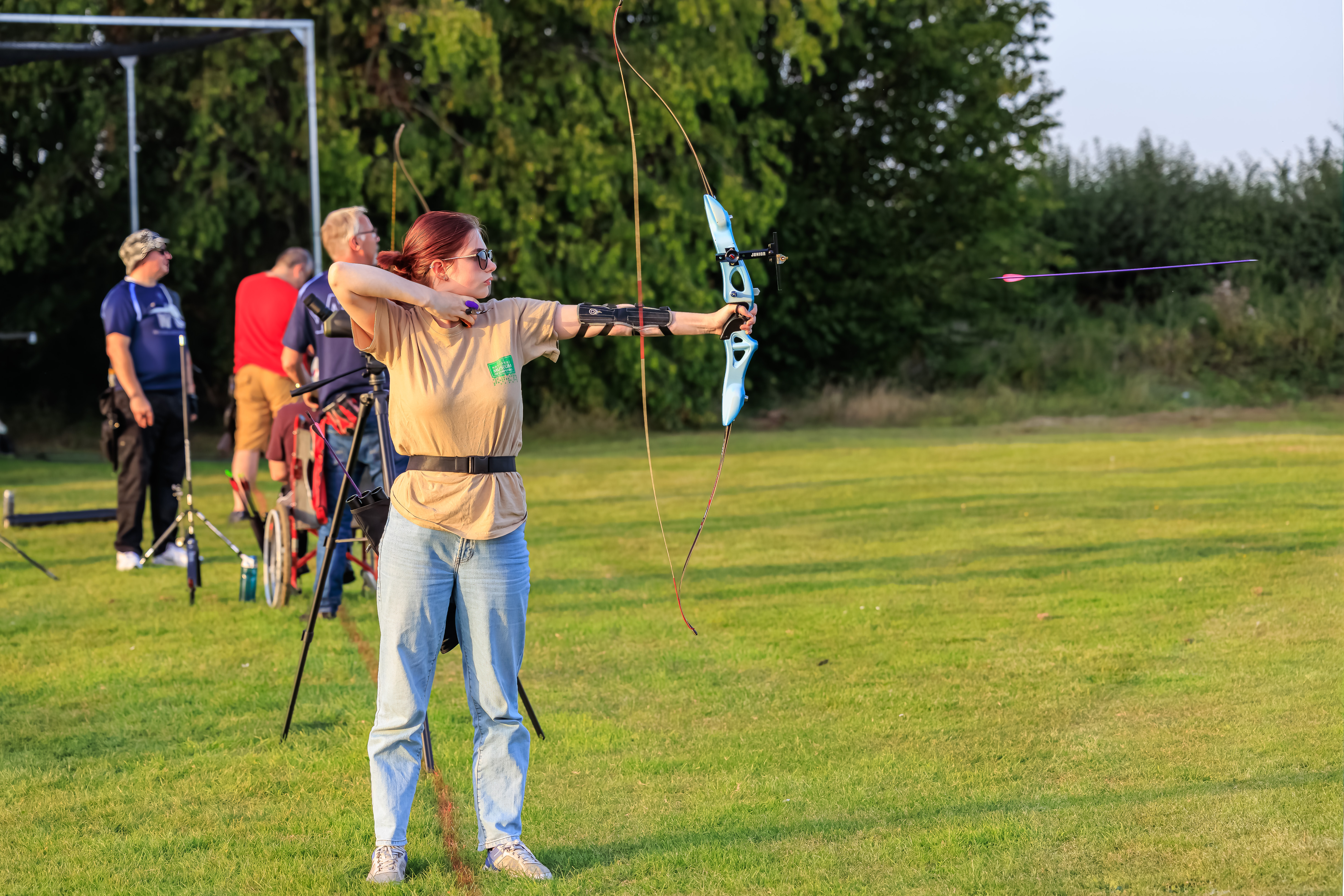 Archers on shooting line, young lady in foreground with auburn hair, sunglasses on has just released and arrow caught in flight after just leaving bow.
